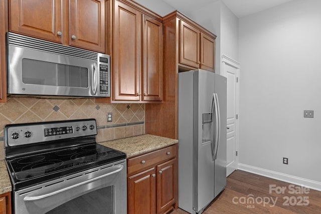 kitchen featuring appliances with stainless steel finishes, wood finished floors, brown cabinets, light stone countertops, and decorative backsplash