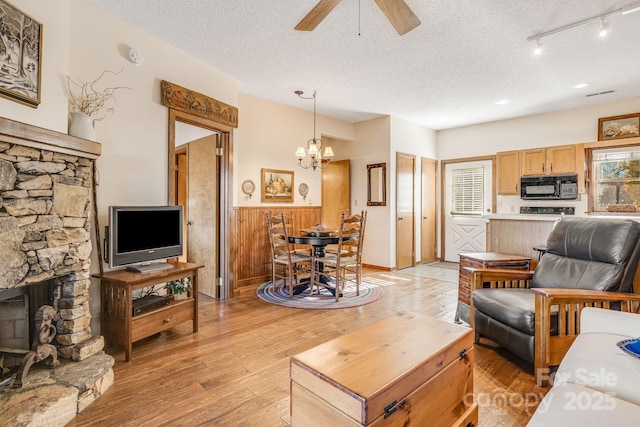 living room with a stone fireplace, wood walls, a textured ceiling, light hardwood / wood-style floors, and ceiling fan with notable chandelier