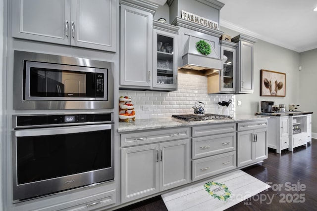 kitchen featuring tasteful backsplash, gray cabinetry, appliances with stainless steel finishes, glass insert cabinets, and light stone countertops