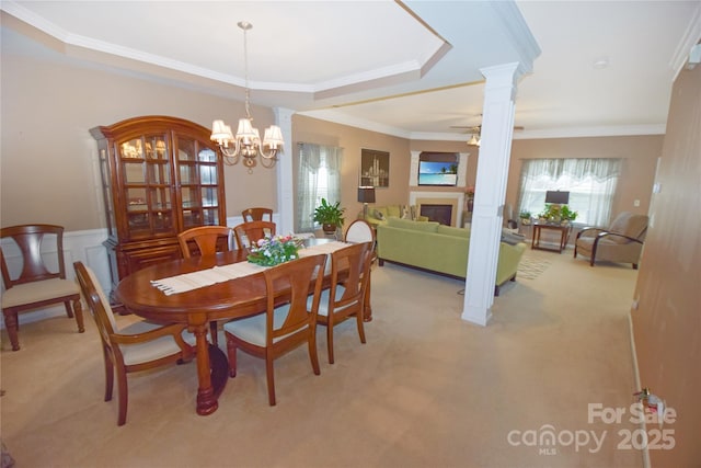 carpeted dining area featuring ceiling fan with notable chandelier, ornamental molding, a raised ceiling, and ornate columns