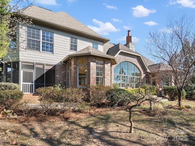 back of property featuring a shingled roof, a sunroom, brick siding, and a chimney