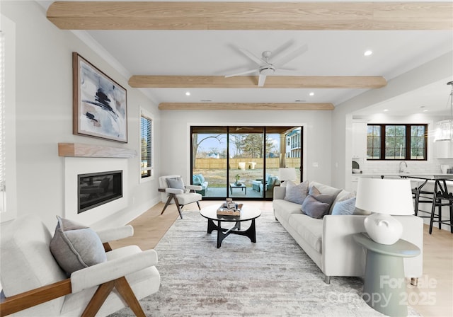 living room featuring light wood finished floors, ceiling fan, beamed ceiling, and a glass covered fireplace