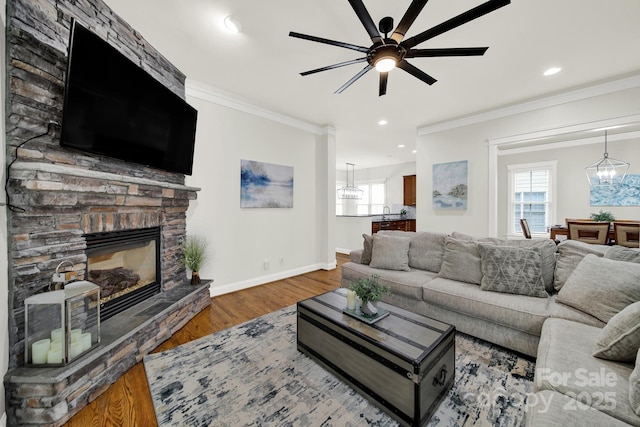 living area with wood finished floors, ornamental molding, baseboards, ceiling fan with notable chandelier, and a stone fireplace