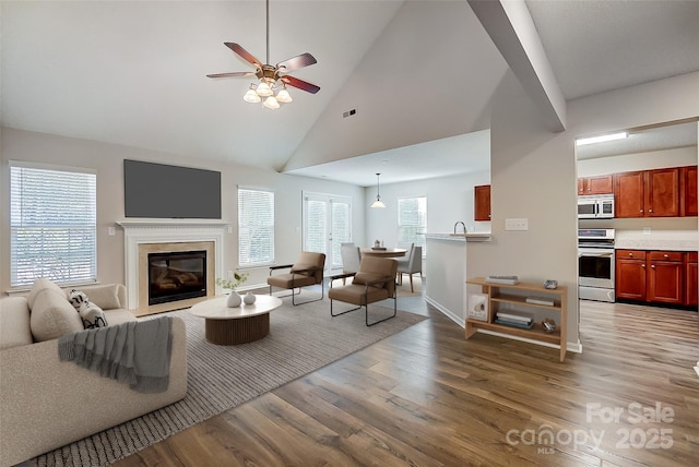 living area with light wood-style floors, a glass covered fireplace, ceiling fan, high vaulted ceiling, and baseboards