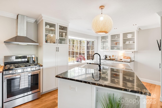 kitchen featuring stainless steel gas range oven, white cabinets, wall chimney exhaust hood, glass insert cabinets, and dark stone countertops