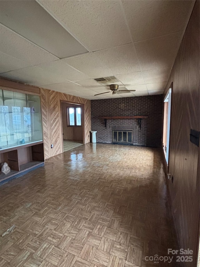 unfurnished living room featuring wood walls, a paneled ceiling, a brick fireplace, parquet floors, and ceiling fan