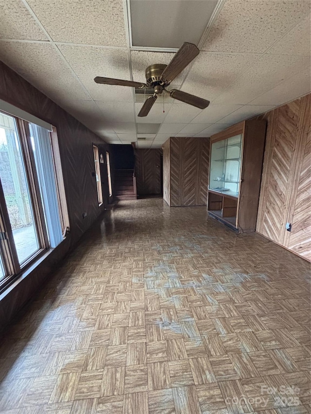 unfurnished living room featuring light parquet floors, ceiling fan, a drop ceiling, and wooden walls