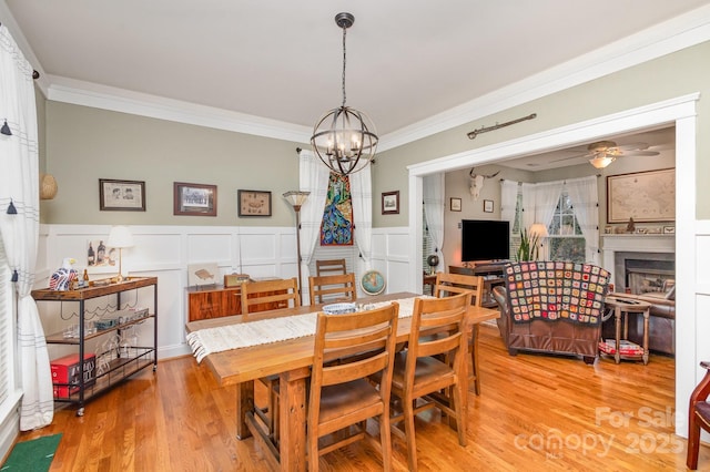 dining space featuring ceiling fan with notable chandelier, a fireplace, ornamental molding, wainscoting, and light wood finished floors