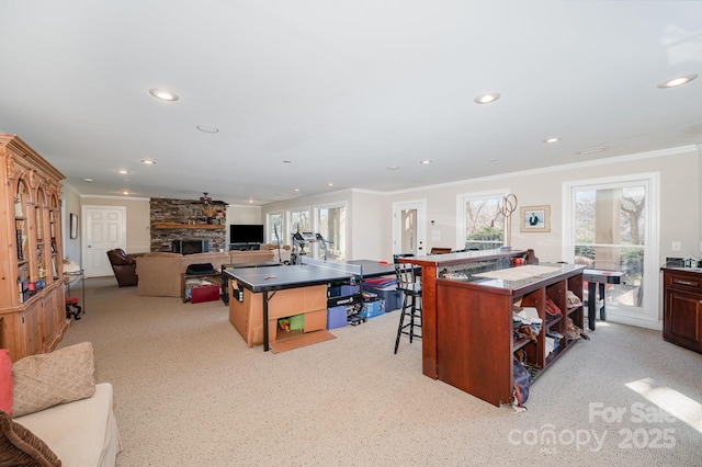 kitchen featuring a kitchen island with sink, light colored carpet, a stone fireplace, ornamental molding, and a kitchen bar