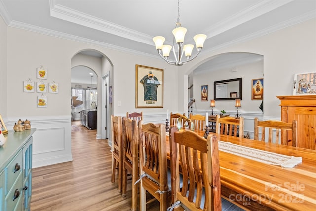 dining area with arched walkways, a tray ceiling, a wainscoted wall, light wood finished floors, and an inviting chandelier