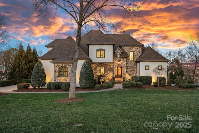 french country style house with stone siding, roof with shingles, and a yard