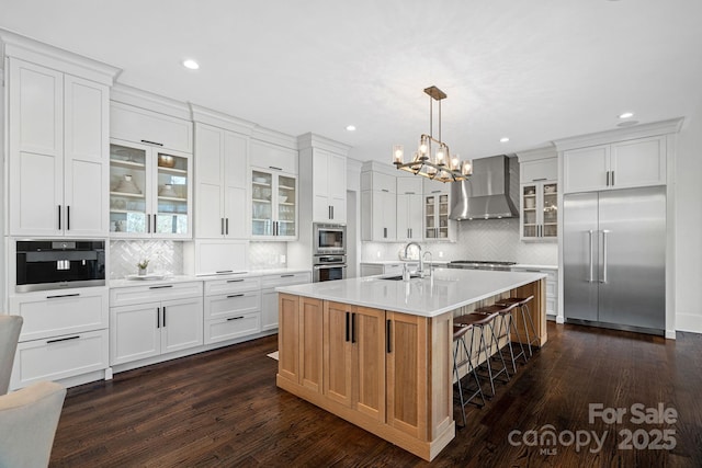 kitchen featuring a center island with sink, wall chimney exhaust hood, glass insert cabinets, built in appliances, and light countertops