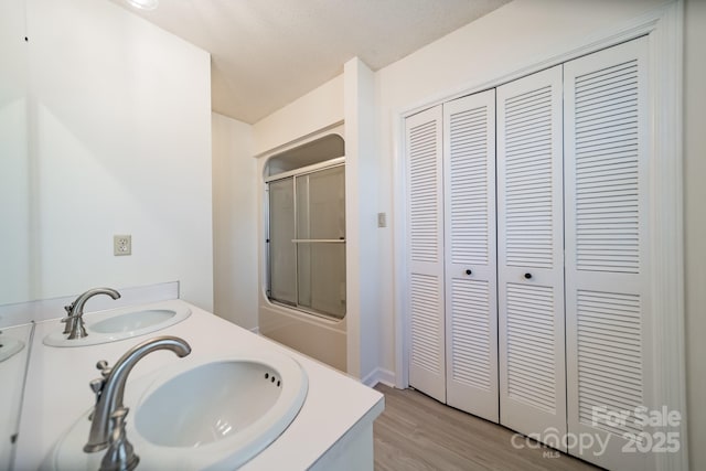 bathroom featuring double vanity, shower / bath combination with glass door, a sink, and wood finished floors