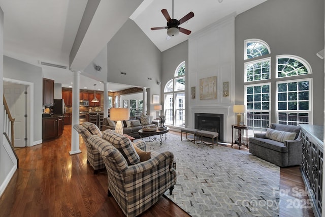 living room featuring decorative columns, a fireplace, a high ceiling, ceiling fan, and dark wood-type flooring