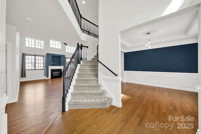 stairway featuring a glass covered fireplace, wood finished floors, a wainscoted wall, and a towering ceiling