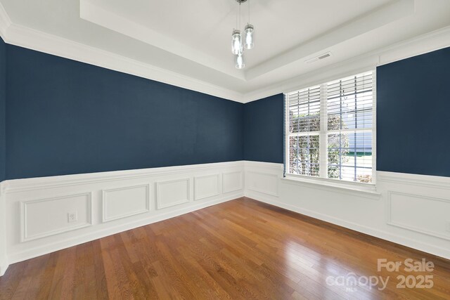 empty room with visible vents, a raised ceiling, wood finished floors, and wainscoting