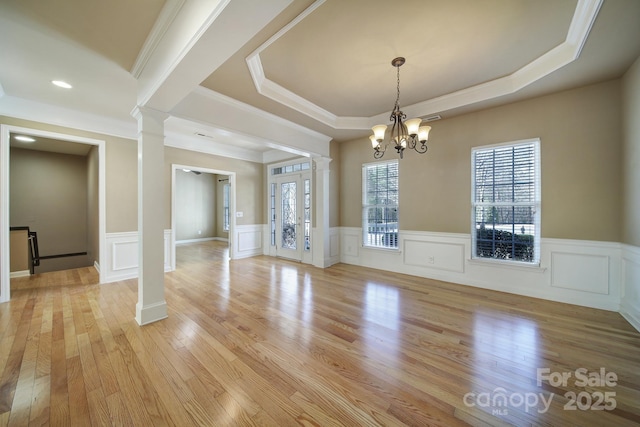 unfurnished dining area with a tray ceiling, light wood-style flooring, an inviting chandelier, wainscoting, and ornate columns