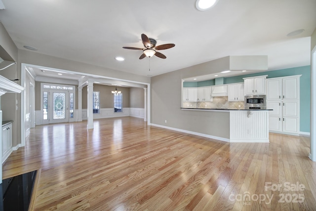 unfurnished living room featuring light wood-style floors, a wainscoted wall, ceiling fan with notable chandelier, ornate columns, and recessed lighting