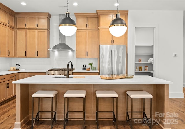 kitchen featuring decorative light fixtures, dark wood finished floors, high end fridge, an island with sink, and wall chimney exhaust hood