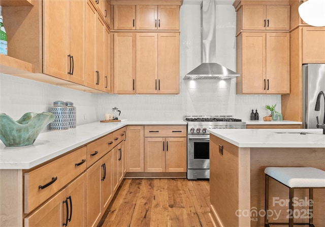 kitchen featuring tasteful backsplash, wall chimney exhaust hood, appliances with stainless steel finishes, light countertops, and light wood-type flooring