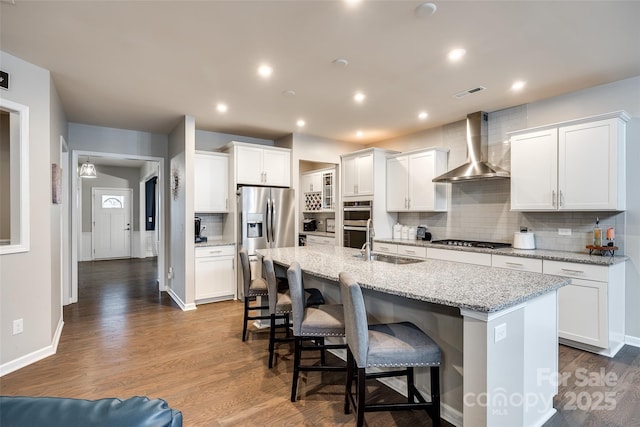 kitchen featuring wall chimney exhaust hood, a center island with sink, white cabinetry, and stainless steel appliances