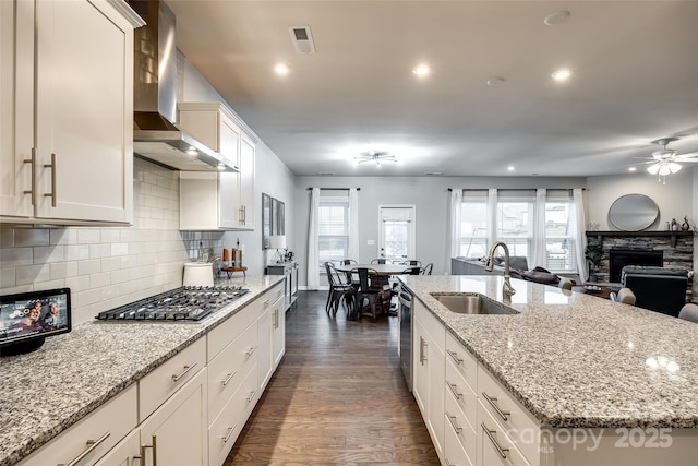 kitchen with light stone counters, a kitchen island with sink, stainless steel appliances, a sink, and wall chimney range hood