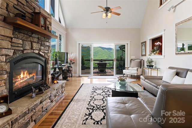 living area featuring a towering ceiling, light wood finished floors, a ceiling fan, and a stone fireplace