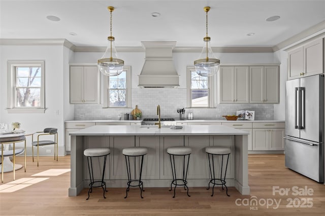 kitchen featuring custom exhaust hood, high end refrigerator, decorative light fixtures, and an island with sink