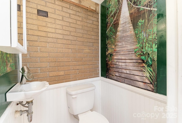 half bath featuring toilet, brick wall, a wainscoted wall, and a sink