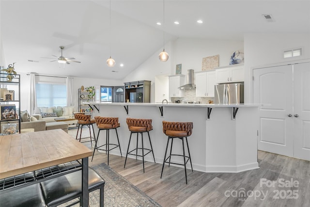 kitchen featuring pendant lighting, wall chimney range hood, stainless steel fridge, a kitchen breakfast bar, and white cabinets