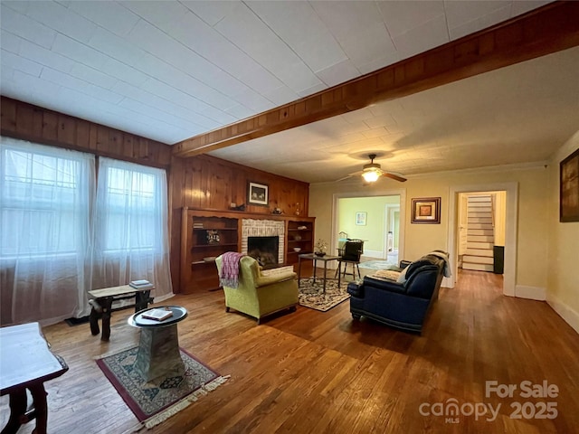 living room featuring ceiling fan, beam ceiling, wood-type flooring, a brick fireplace, and wood walls