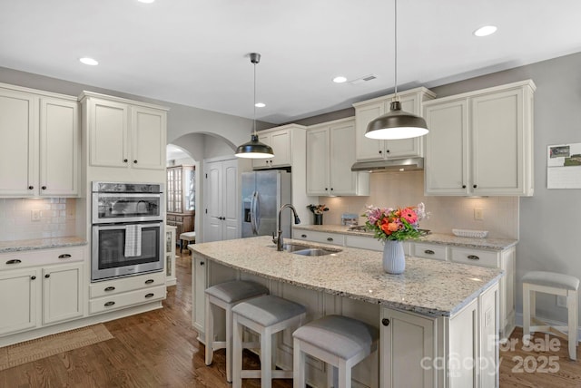 kitchen with dark wood-type flooring, under cabinet range hood, appliances with stainless steel finishes, arched walkways, and a sink