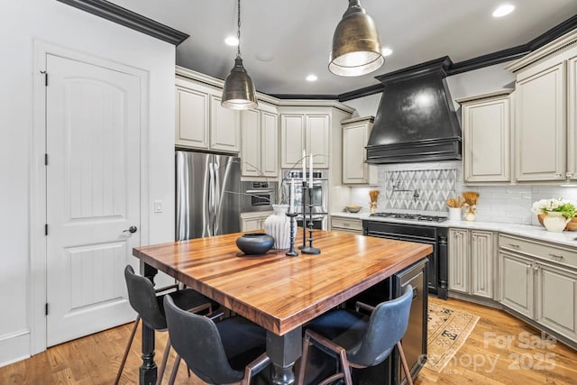 kitchen with appliances with stainless steel finishes, hanging light fixtures, butcher block counters, custom exhaust hood, and a breakfast bar area