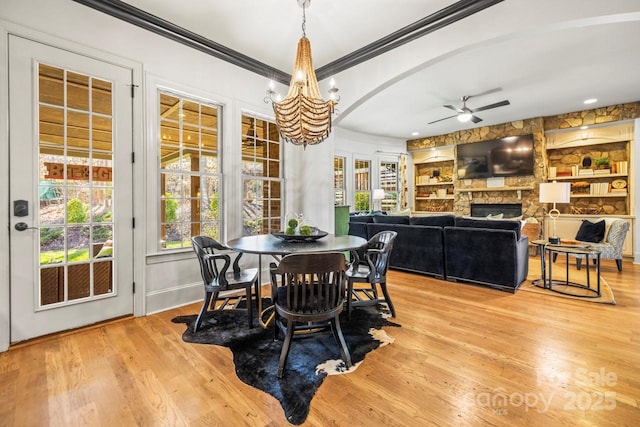 dining space featuring built in shelves, a stone fireplace, ceiling fan with notable chandelier, light hardwood / wood-style floors, and ornamental molding