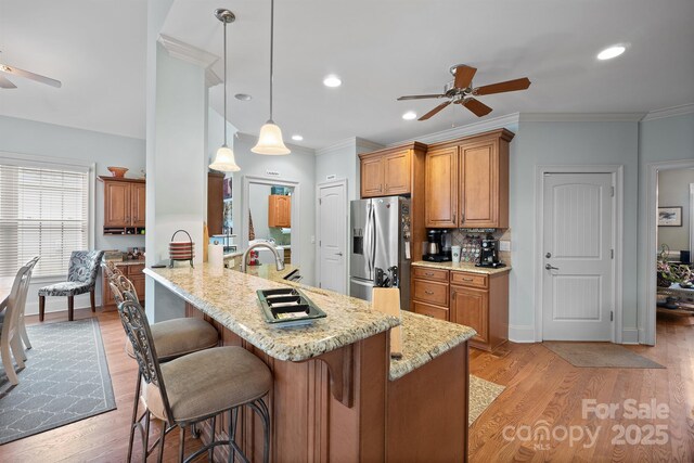 kitchen featuring ceiling fan, brown cabinetry, a breakfast bar area, stainless steel fridge with ice dispenser, and light stone counters