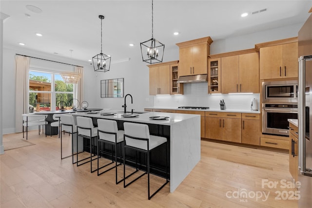 kitchen with glass insert cabinets, appliances with stainless steel finishes, a kitchen island with sink, under cabinet range hood, and light brown cabinets