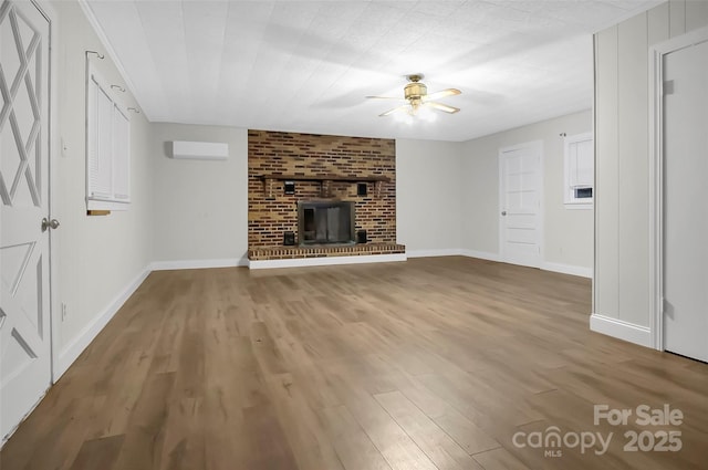 unfurnished living room featuring a fireplace, an AC wall unit, ceiling fan, wood finished floors, and baseboards