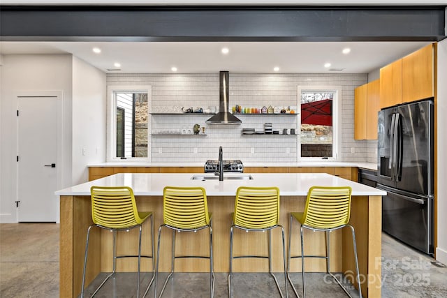 kitchen with black fridge with ice dispenser, a kitchen breakfast bar, a kitchen island with sink, light countertops, and open shelves