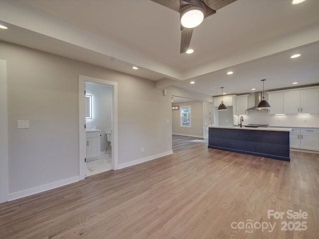 kitchen featuring white cabinetry, a center island with sink, light wood-type flooring, pendant lighting, and wall chimney range hood