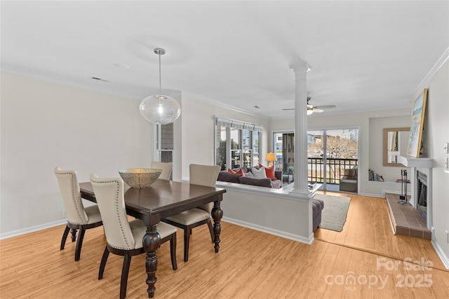 dining area with light wood finished floors, decorative columns, a fireplace with raised hearth, a ceiling fan, and crown molding