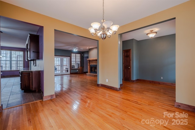 unfurnished dining area with light wood finished floors, baseboards, a fireplace with raised hearth, french doors, and ceiling fan with notable chandelier