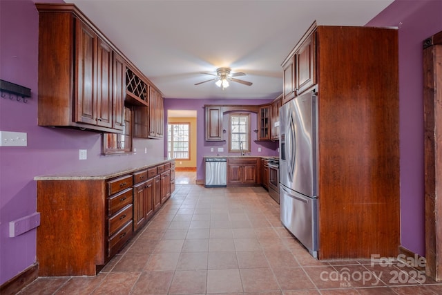 kitchen featuring light tile patterned floors, stainless steel appliances, a ceiling fan, brown cabinets, and glass insert cabinets