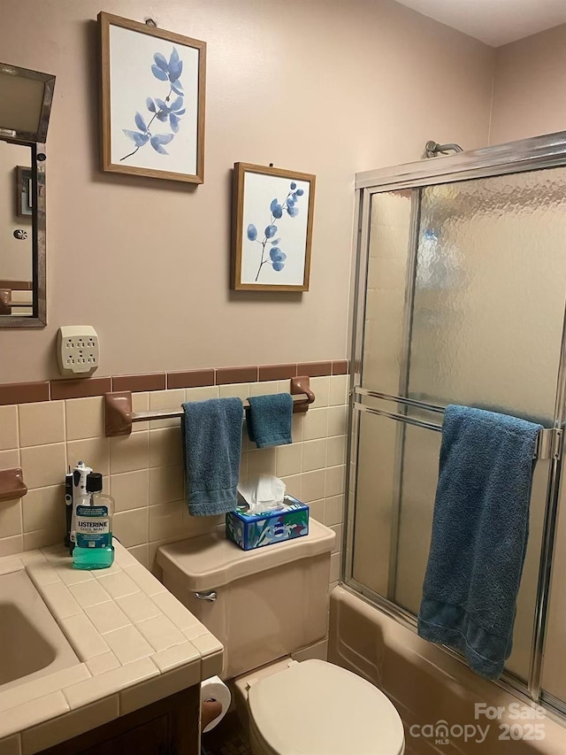 full bath with wainscoting, toilet, combined bath / shower with glass door, vanity, and tile walls