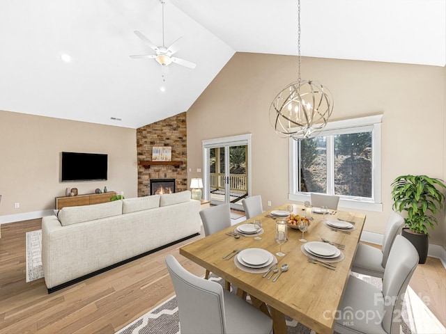 dining area with a stone fireplace, ceiling fan with notable chandelier, high vaulted ceiling, and light wood-type flooring