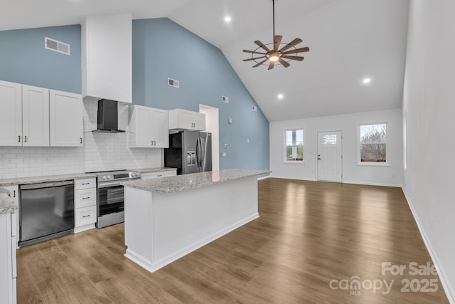 kitchen featuring light stone counters, appliances with stainless steel finishes, wall chimney range hood, and white cabinetry