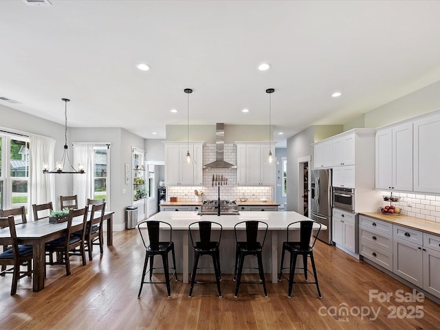 kitchen featuring decorative light fixtures, white cabinetry, a kitchen island with sink, stainless steel appliances, and wall chimney range hood