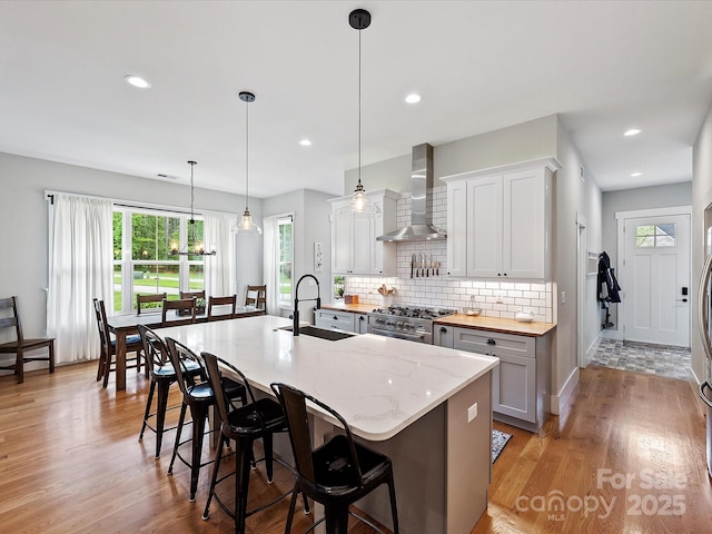 kitchen featuring a kitchen bar, wall chimney exhaust hood, decorative light fixtures, a kitchen island with sink, and white cabinets