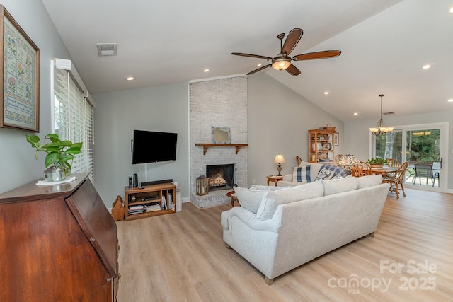 living room featuring ceiling fan with notable chandelier, a brick fireplace, visible vents, light wood finished floors, and lofted ceiling