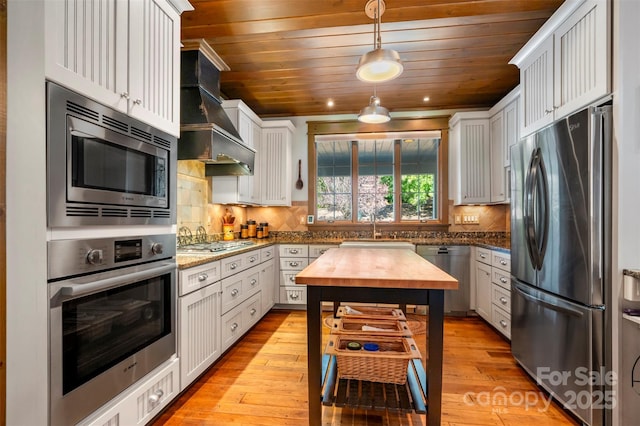 kitchen featuring appliances with stainless steel finishes, white cabinetry, hanging light fixtures, light stone counters, and custom range hood