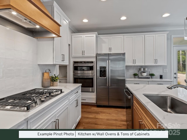 kitchen featuring wood finished floors, custom exhaust hood, a sink, stainless steel appliances, and white cabinetry